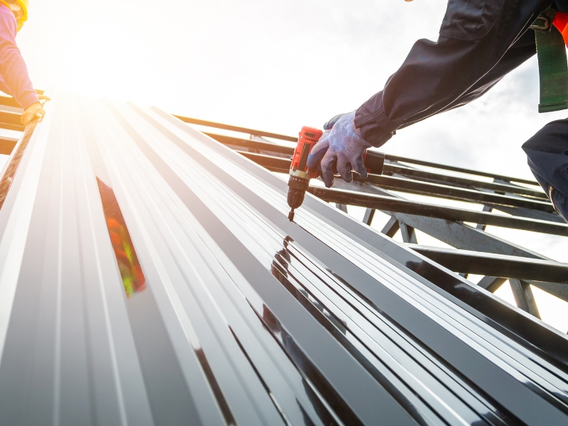 Worker using a drill while installing sheet metal roofing on a high sloped structure.