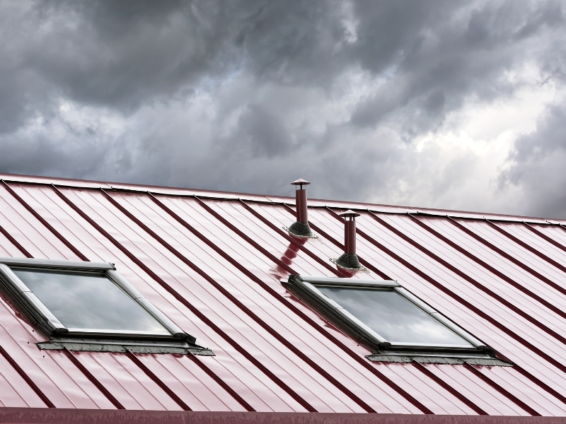 Installing a skylight on red metal roofing with vent pipes and stormy skies in the background